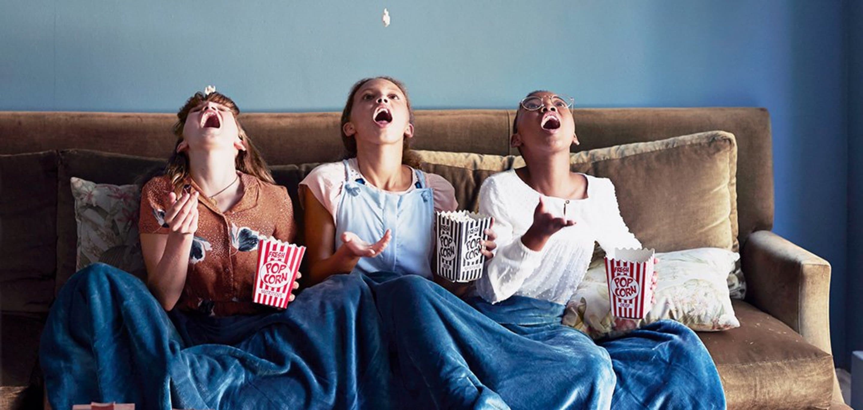 Kabelfernsehen HD-Pakete Drei Mädchen auf einer Couch werfen Popcorn in die Luft, und versuchen es mit dem Mund aufzufangen