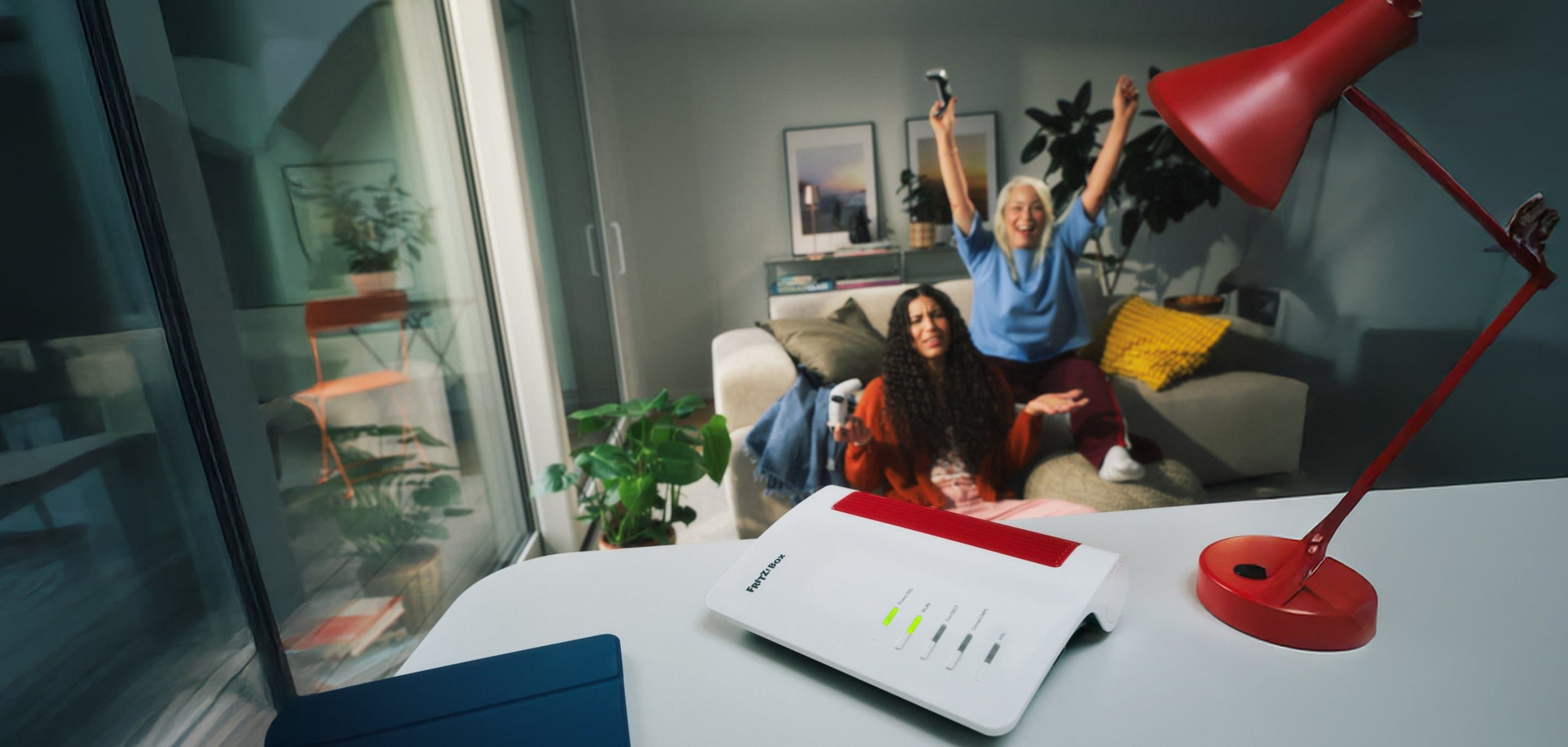 Im Vordergrund steht ein Router auf einem Tisch; im Hintergrund sitzen zwei Personen auf einem Sofa und reagieren auf ein Geschehen außerhalb des Bildausschnitts.