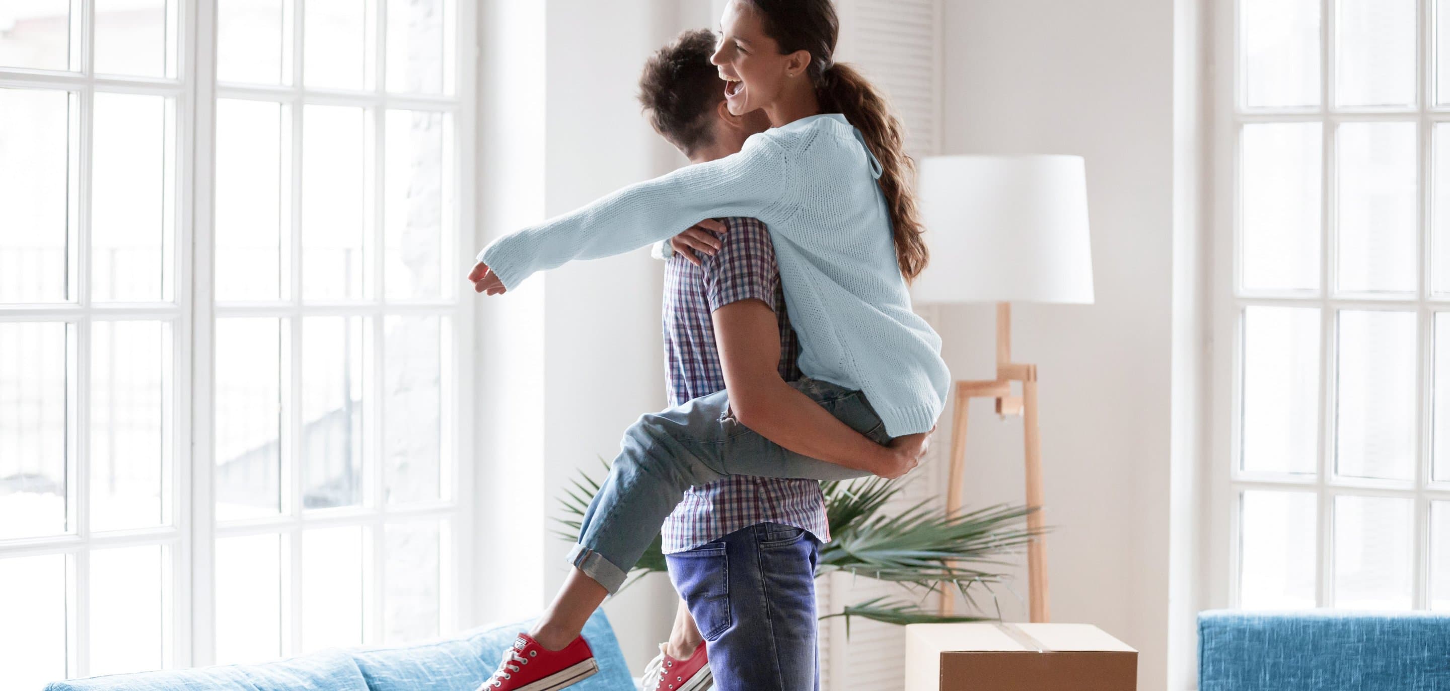 Mann hebt lachende Frau hoch. Im Hintergrund sind Umzugskartons zu sehen.