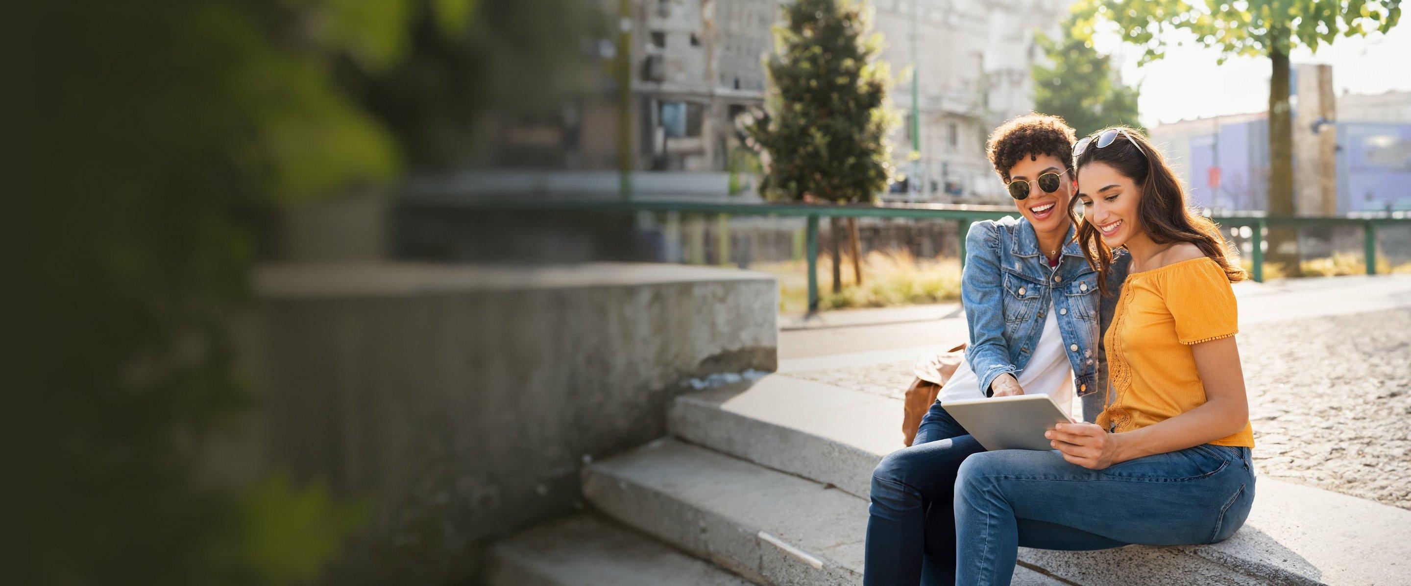 Zwei Frauen sitzen draußen auf einer Treppe und schauen auf den Laptop