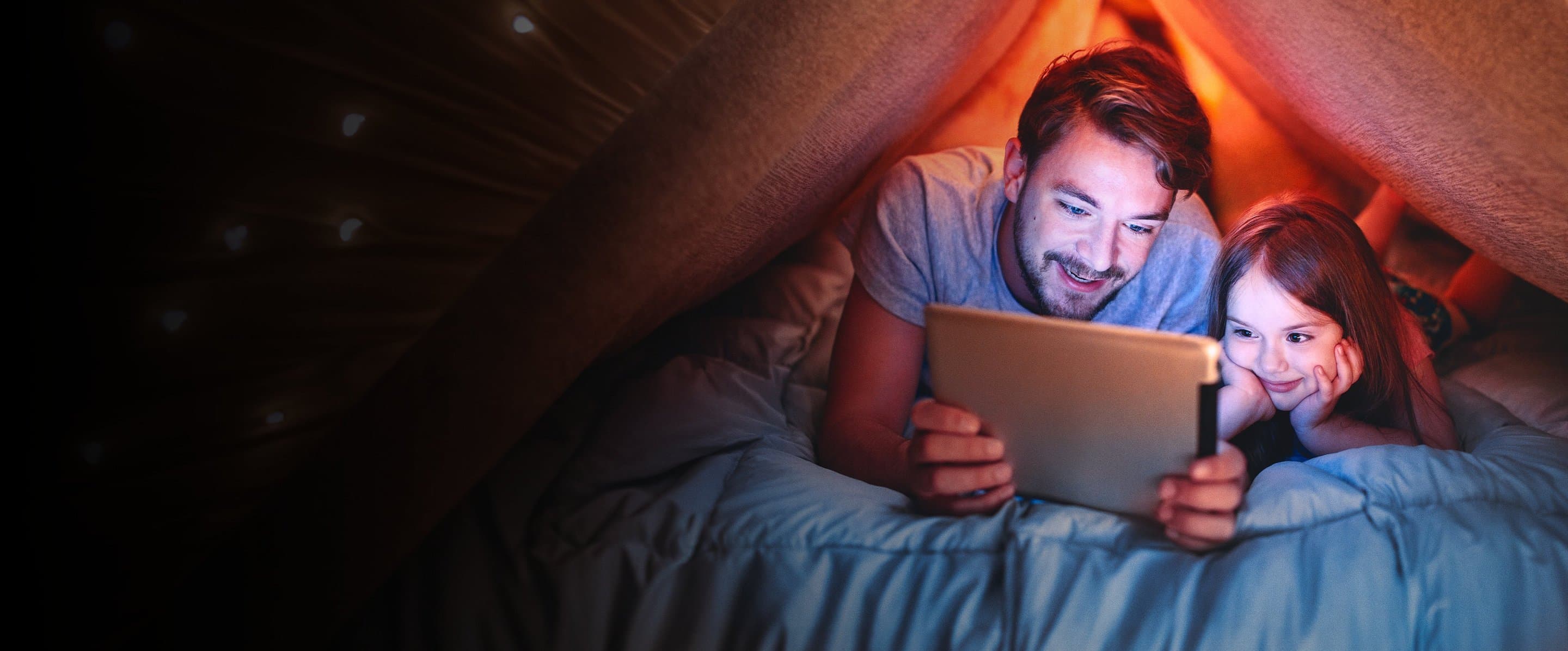 Ein Vater und seine Tochter liegen gemeinsam unter einer Decke und schauen auf ein Tablet.