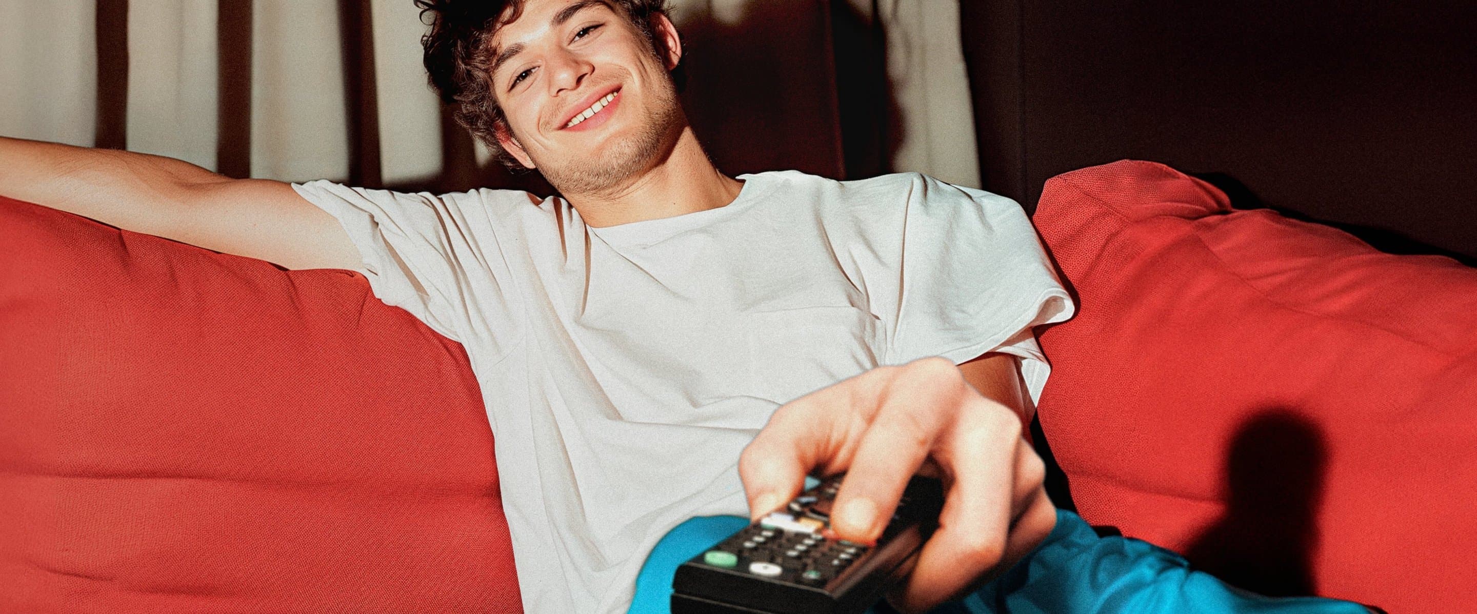 NetTV Verfügbarkeit prüfen Junger Erwachsener sitzt auf der Couch und hält eine Fernbedienung in der Hand
